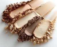 Crushed makeup powder swatches in nude and brown tones with scattered particles showing texture and pigmentation on white background for cosmetic presentation.