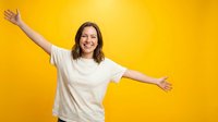 Woman with shoulder-length brown hair, Caucasian ethnicity, wearing white t-shirt with arms outstretched in celebratory pose against bright yellow background, expressing joy and freedom.