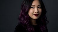 Young Asian woman with purple ombre hair and dark lipstick smiling confidently against dark background. Professional headshot for business and lifestyle.