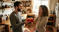Young Caucasian man surprising woman with red gift box in cozy kitchen during romantic moment together at home.