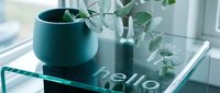 Teal ceramic pot with eucalyptus branches on a glass table with hello inscription, cozy modern interior decor near a bright window.