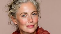 Elegant mature woman with silver hair and blue eyes wearing rust-colored sweater against neutral beige background, showcasing natural beauty and confidence.