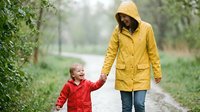 Mother and child walking together in yellow and red raincoats during rainy weather on park path. Family outdoor activity in spring or autumn season.