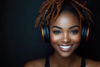 Young African American woman with dreadlocks and bright smile wearing wireless headphones against dark background, radiating joy and confidence in studio portrait.