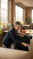 Two male colleagues collaborating at laptop in modern loft office with brick walls and natural light from large windows, suitable for business, teamwork, and corporate projects.