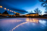 Outdoor ice skating rink at dusk with string lights creating magical atmosphere, silhouettes of people skating, and dramatic blue winter sky reflecting on glossy ice surface.