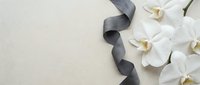 White orchid flowers and dark gray ribbon on beige stone background, flat lay with copy space for funeral or mourning design.