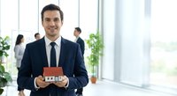 Professional Hispanic businessman in dark suit holding miniature house model in modern office environment for real estate and property investment concepts.