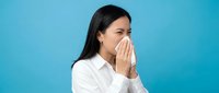 Young woman with dark hair and South Asian ethnicity wearing white shirt sneezing or coughing into her hand against blue background, depicting illness, cold, flu, or allergy symptoms.