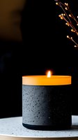 Minimalist black concrete candle holder with orange wax and burning flame against dark background, decorated with delicate dried branches. Perfect for spa and wellness.