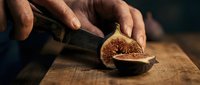 Male hands slicing fresh fig in half on wooden cutting board, showing interior seeds and flesh for culinary preparation and food styling.