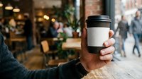 Male hand holding reusable coffee cup with blank label in urban street setting. Eco-friendly beverage container for branding mockups and sustainable lifestyle marketing campaigns.