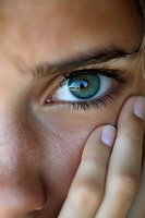 Close-up of bright green eye with long eyelashes and natural freckles on young woman's face, hand partially covering view, soft natural lighting creates intimate mood.