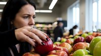 Middle-aged Caucasian woman with dark hair selecting fresh red apple in grocery store produce section with colorful fruits display in background.