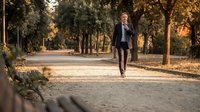 Caucasian male professional in dark business suit walking alone on tree-lined park path during golden hour, suitable for corporate, lifestyle, and business concepts.