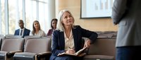 Professional blonde businesswoman taking notes during corporate meeting in modern conference room with colleagues in background.