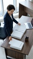 Asian female professional in dark blazer reviewing documents at wooden desk with laptop, overhead view for business and office work concepts.