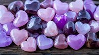 Collection of polished heart-shaped gemstones in various shades of purple, pink, and rose quartz on rustic wooden surface. Natural crystal healing stones.