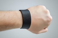 Male wrist wearing wide black leather bracelet or wristband against light gray background, closeup view showing texture and minimalist style.