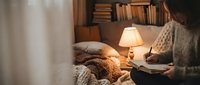 Cozy bedroom scene with person reading book in warm lamplight, soft blankets and pillows creating comfortable atmosphere for relaxation and leisure reading.