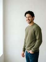 Young man with dark curly hair and olive skin tone wearing sage green sweatshirt and blue jeans, smiling at camera in studio setting with white background for portrait and lifestyle use.