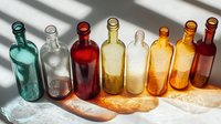 Colorful glass bottles in row casting shadows on white surface. Transparent empty containers in turquoise, clear, red, amber and orange tones create artistic light pattern.