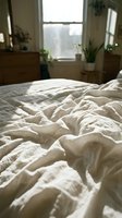 Unmade bed with cream linen sheets in bright bedroom with large window, wooden furniture, and potted plants creating serene morning atmosphere for lifestyle and home decor content.