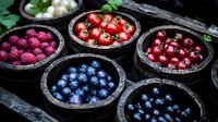 Fresh berries and fruits in rustic wooden bowls on dark background, including blueberries, raspberries, cherries and tomatoes, arranged in moody food composition.