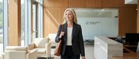 Professional blonde businesswoman in black suit standing confidently in modern corporate office lobby with natural lighting and contemporary furniture.