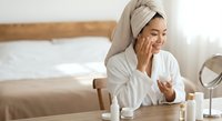 Young Asian woman in white bathrobe and towel applying skincare cream in modern bedroom with natural sunlight for morning beauty routine and self-care.