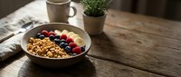 Granola bowl with fresh berries and banana slices on wooden table, styled with coffee mug and potted plant for breakfast or healthy eating content.