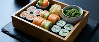 Assorted sushi set in wooden bento box with salmon rolls, maki, pickled ginger and seaweed salad on dark slate background.