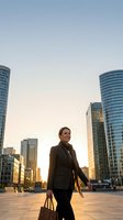 Businesswoman in dark blazer holding leather briefcase standing in modern urban plaza surrounded by tall glass skyscrapers at golden hour sunrise, corporate professional environment for business and finance concepts.