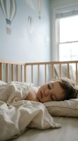 Peaceful toddler boy sleeping in wooden crib with soft white bedding in bright nursery room with hot air balloon wall decor and natural window light.