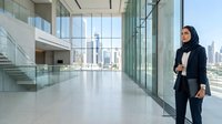 Woman in professional business attire wearing hijab standing in modern office lobby with floor-to-ceiling windows overlooking city skyline, suitable for corporate and diversity concepts.