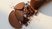 Broken bronze powder compact with makeup brush and scattered particles on neutral background, suitable for beauty and cosmetics advertising.