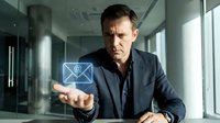 Mature male professional with dark hair and light skin tone wearing navy blazer holds glowing email icon in modern office setting with glass walls and natural light for business communication concepts.