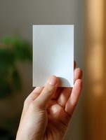 Female hand holding blank white paper card against blurred natural background, mockup for business card or invitation design presentation.