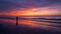 Solitary figure walking on wet sand beach during dramatic sunset with vibrant purple and orange sky reflecting on water creating peaceful coastal scene.