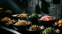 Assorted Asian noodle dishes and fresh ingredients displayed in black bowls on dark surface, featuring stir-fried noodles, vegetables, and cured meats for restaurant, menu, or food styling.