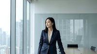 Professional Asian businesswoman in navy suit standing by office windows overlooking city skyline, corporate executive portrait for business presentations.