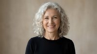 Confident mature Caucasian woman with curly gray hair smiling warmly in black top against neutral background for professional headshot and lifestyle content.