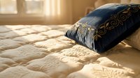 Decorative navy pillow with gold embroidery rests on cream quilted bedding in sunlit bedroom, showcasing luxury home decor and interior design styling.