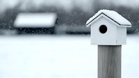 White wooden birdhouse covered in snow against blurred winter background, minimalist composition with soft bokeh effect creates serene seasonal atmosphere.