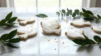 Homemade bird-shaped sugar cookies dusted with powdered sugar arranged on white surface with fresh eucalyptus leaves for baking and holiday decoration.