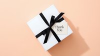 White gift box with black ribbon bow and thank you tag on pastel peach background, top view for holiday or celebration concept.