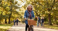 Happy mature Caucasian woman cycling through autumn park with wicker basket, enjoying outdoor recreation and healthy lifestyle during golden fall season.