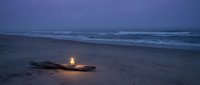 Driftwood campfire glowing on sandy beach at twilight with ocean waves in background creating peaceful coastal evening atmosphere for relaxation and meditation.