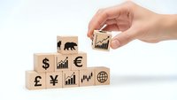 Hand placing wooden block with growth chart onto stack of currency and financial symbol blocks representing global investment strategy.