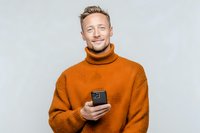 Young Caucasian man with beard wearing orange turtleneck sweater holding smartphone, smiling against light gray background.